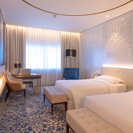 Steigenberger Hotel Doha Exteriör bild The photo shows a modern hotel room with two single beds placed beside each other. The room features a contemporary design with light-colored walls and stylish decor. There is a large window dressed with sheer curtains, allowing natural light to fill