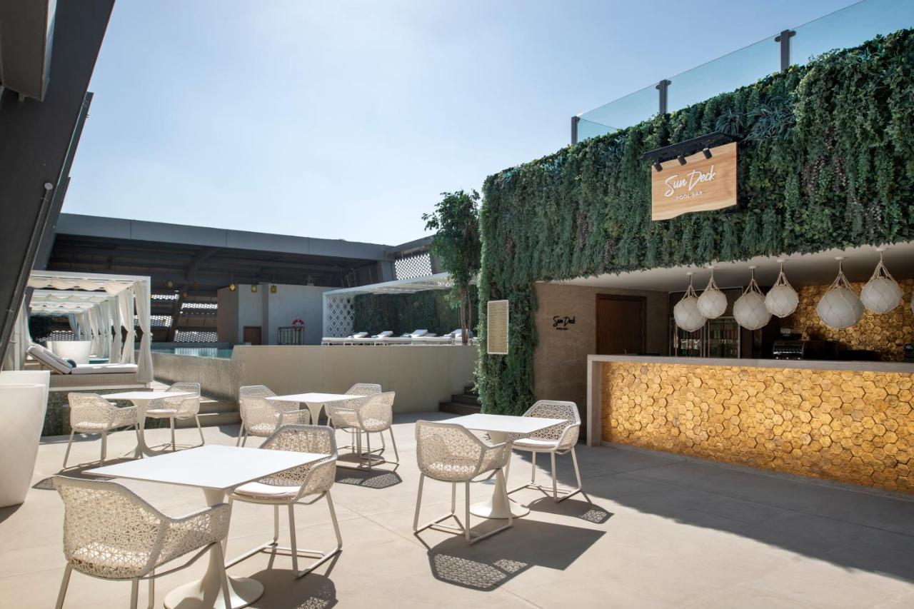 Steigenberger Hotel Doha Exteriör bild The photo shows a stylish outdoor seating area of a modern venue, likely a restaurant or bar. The space features several white tables and chairs arranged neatly on a light-colored floor. In the background, there's a bar area with a warm-toned wood fa