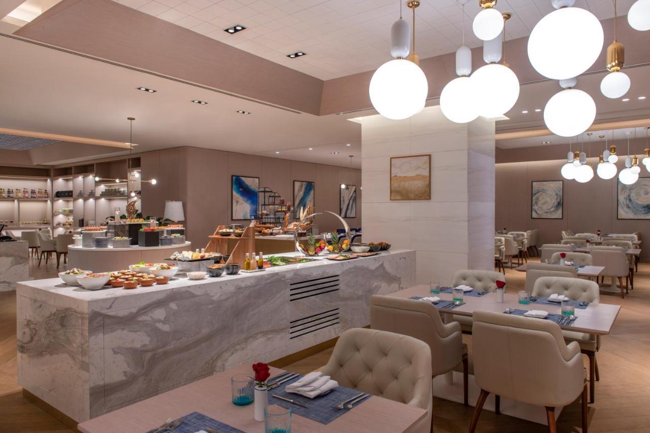 Steigenberger Hotel Doha Exteriör bild The photo shows a modern restaurant interior that features a stylish and elegant design. In the foreground, there are tables with light-colored upholstered chairs, set with blue napkins and small flower arrangements. 

In the background, there is a b