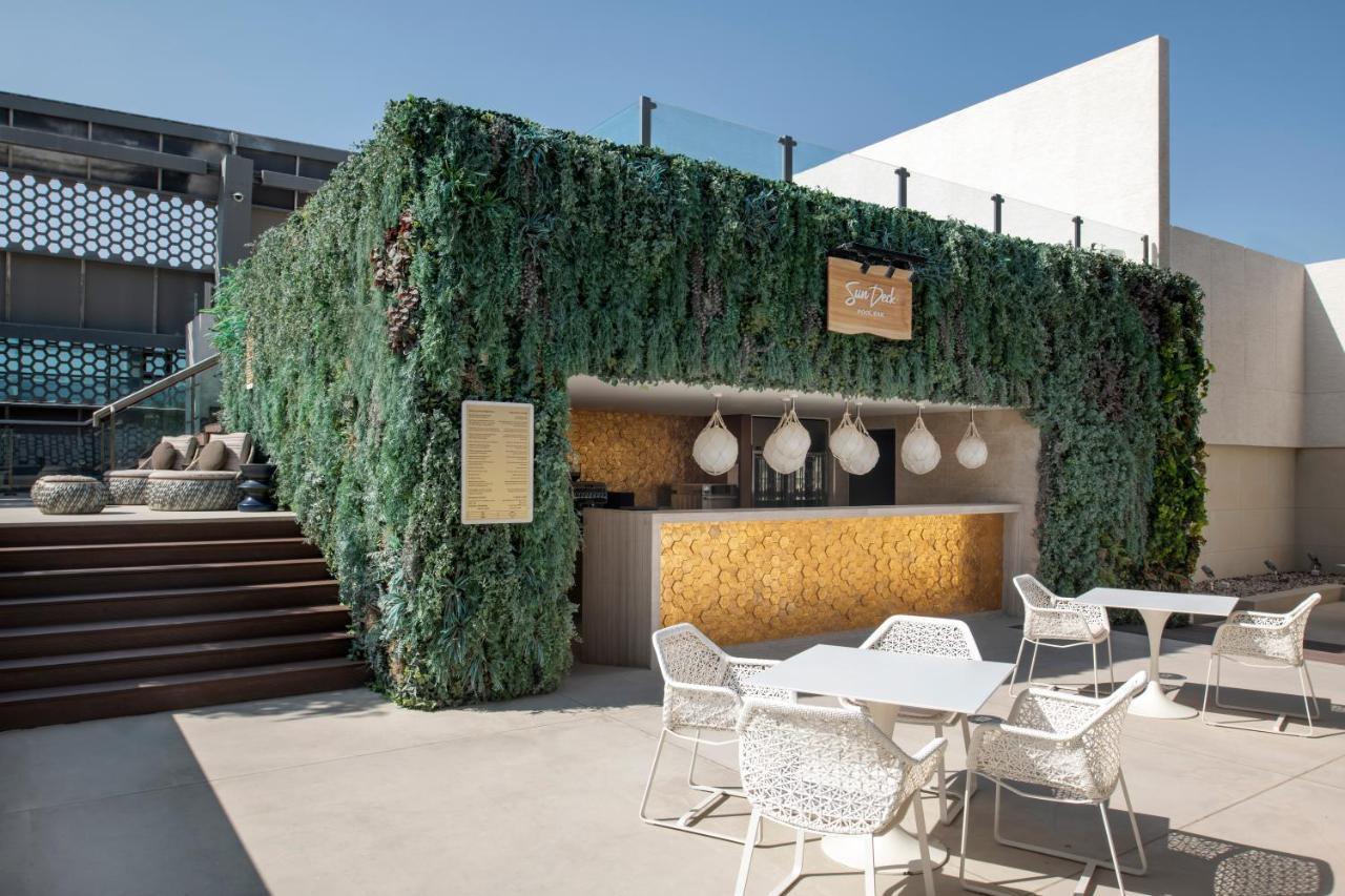 Steigenberger Hotel Doha Exteriör bild The photo shows an outdoor bar area surrounded by lush greenery. The bar has a modern design, featuring a wooden counter with decorative pendant lights hanging above. There are several white outdoor tables and chairs arranged in front of the bar, whi