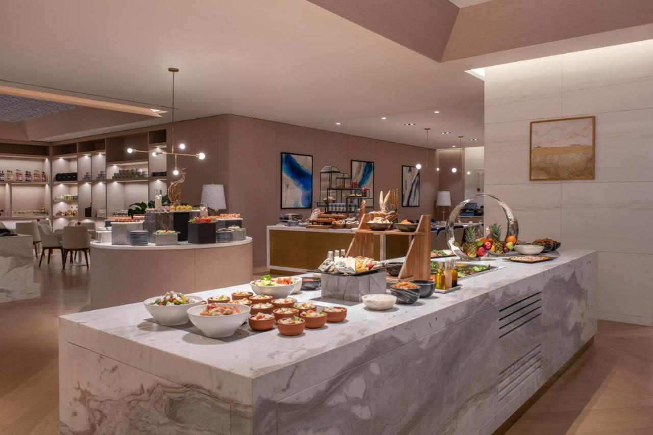 Steigenberger Hotel Doha Exteriör bild The photo depicts a modern dining area, likely a buffet setup in a restaurant or hotel. A long white marble counter is filled with various dishes in bowls and platters, showcasing an assortment of food items. The space has a sophisticated and elegant