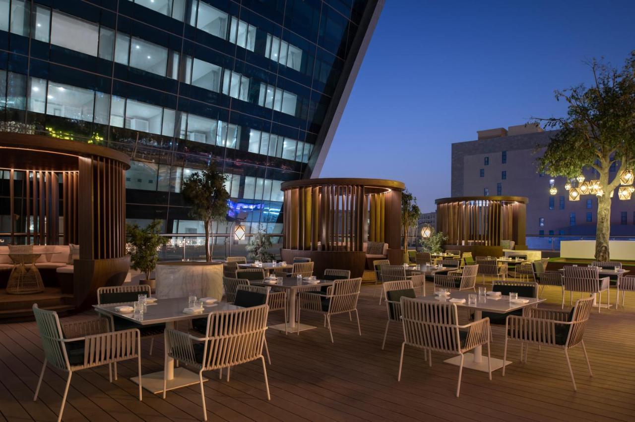 Steigenberger Hotel Doha Exteriör bild The photo shows an outdoor dining area that features several tables and chairs arranged neatly on a wooden deck. The seating includes modern white chairs and a mix of seating styles, including some areas with privacy screens or structures that provid