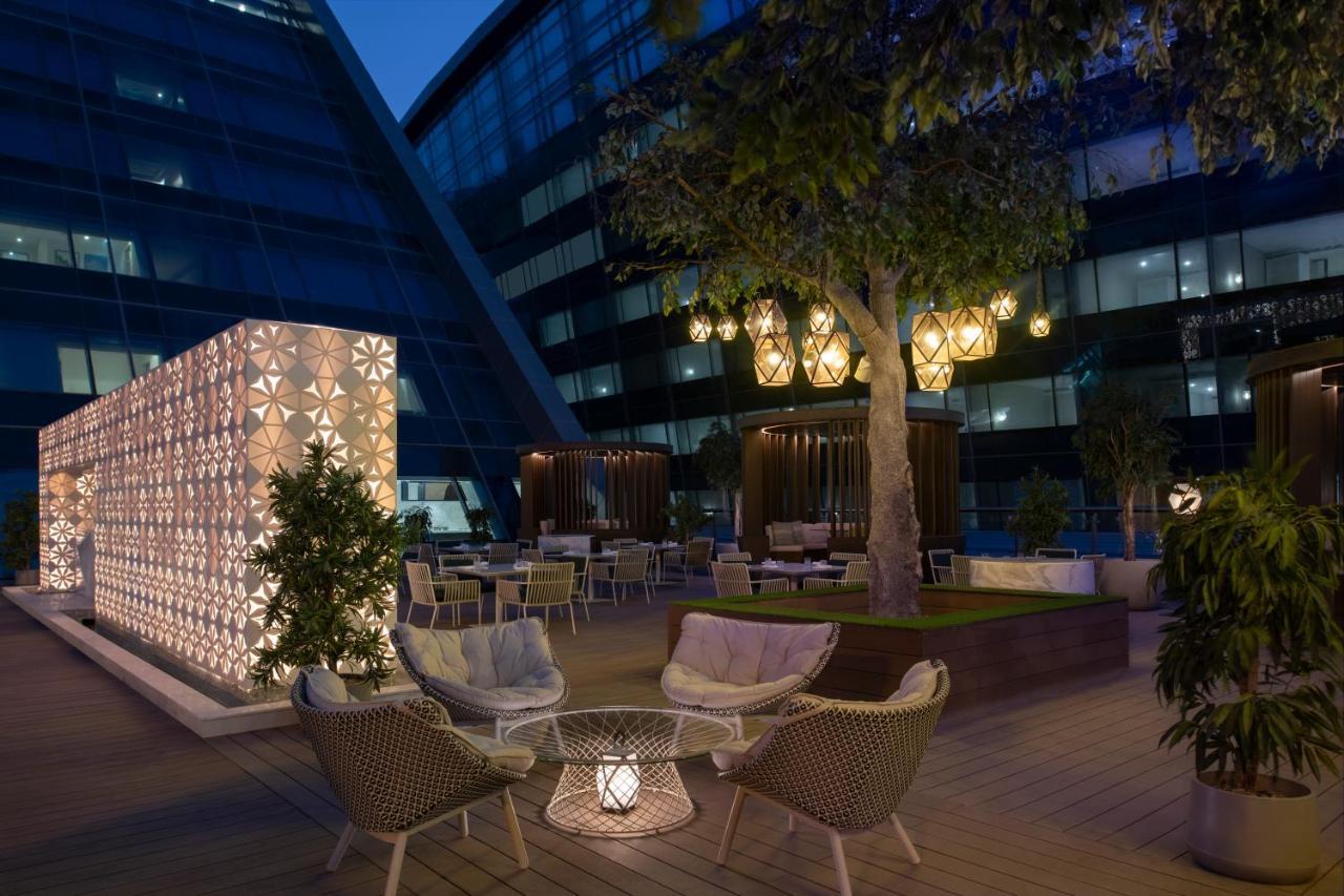 Steigenberger Hotel Doha Exteriör bild The photo depicts a stylish outdoor terrace area at night. There are modern seating arrangements, featuring woven chairs and a decorative table, surrounded by lush greenery. A large tree is present, adding to the ambiance. 

On one side, there's a st