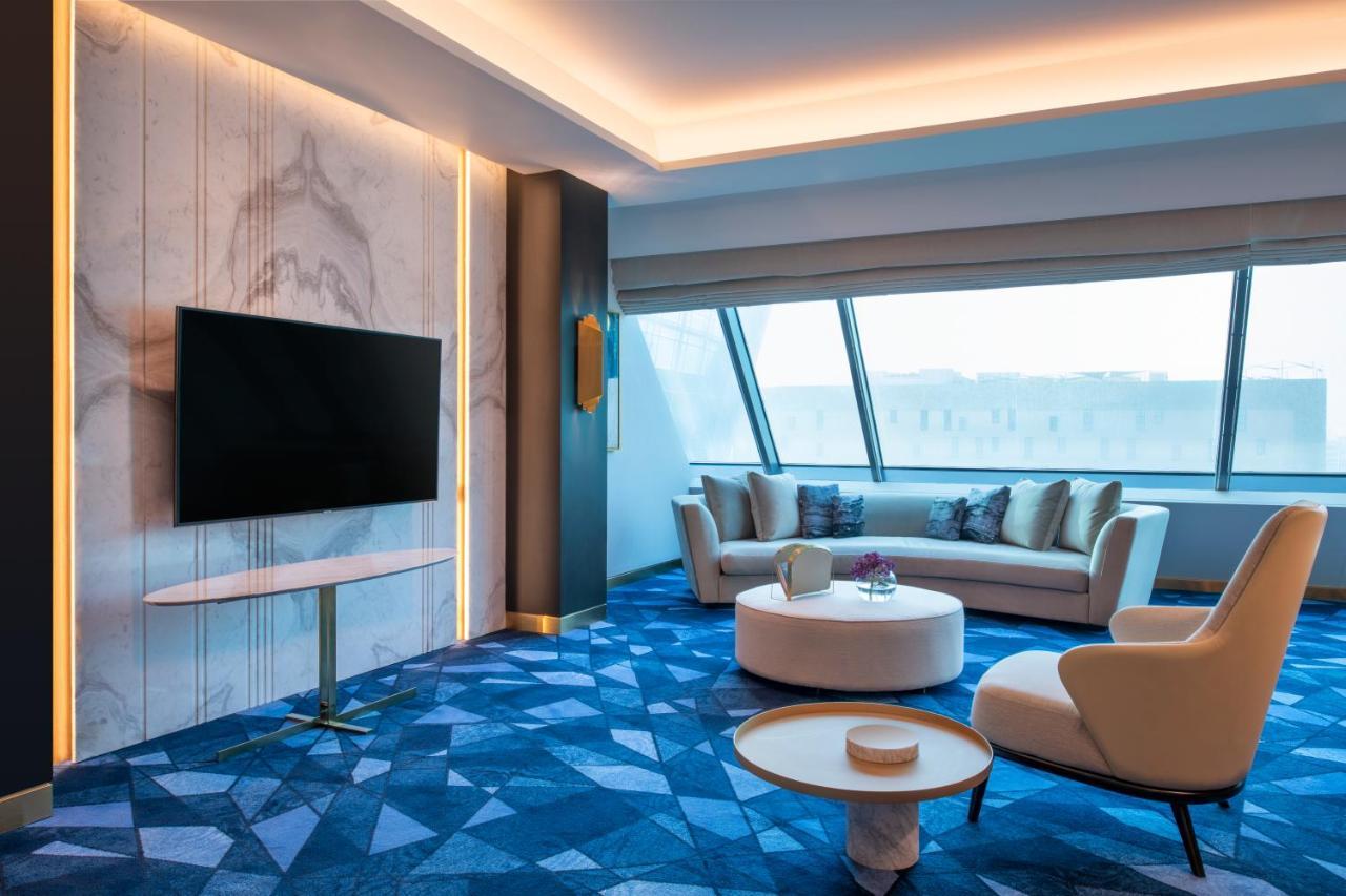 Steigenberger Hotel Doha Exteriör bild The photo shows a modern and stylish living room with a large window that provides plenty of natural light. There is a flat-screen television mounted on a wall, and a comfortable sofa with cushions is positioned nearby. A round coffee table sits in f