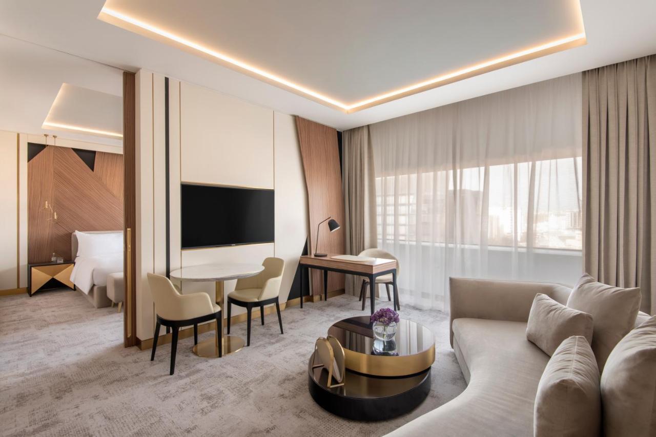 Steigenberger Hotel Doha Exteriör bild The photo shows a modern hotel room that features a stylish and contemporary design. There is a comfortable seating area with a light-colored sofa and a round coffee table, along with two chairs and a small desk. A large flat-screen television is mou