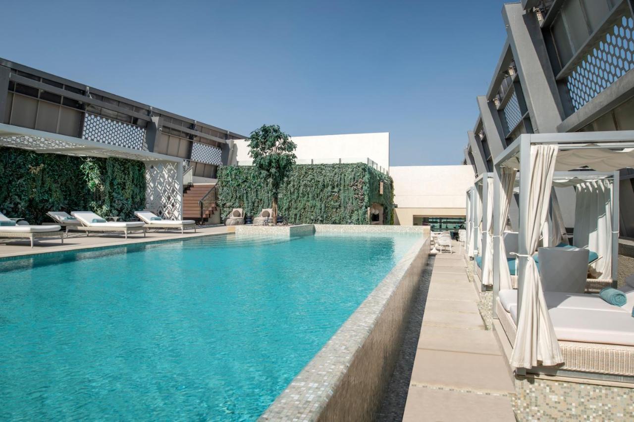Steigenberger Hotel Doha Exteriör bild The image shows a modern, outdoor swimming pool area. The pool is large and has bright blue water, surrounded by a sleek stone deck. There are several lounge chairs positioned beside the pool, some of which are covered by canopies. To one side, there