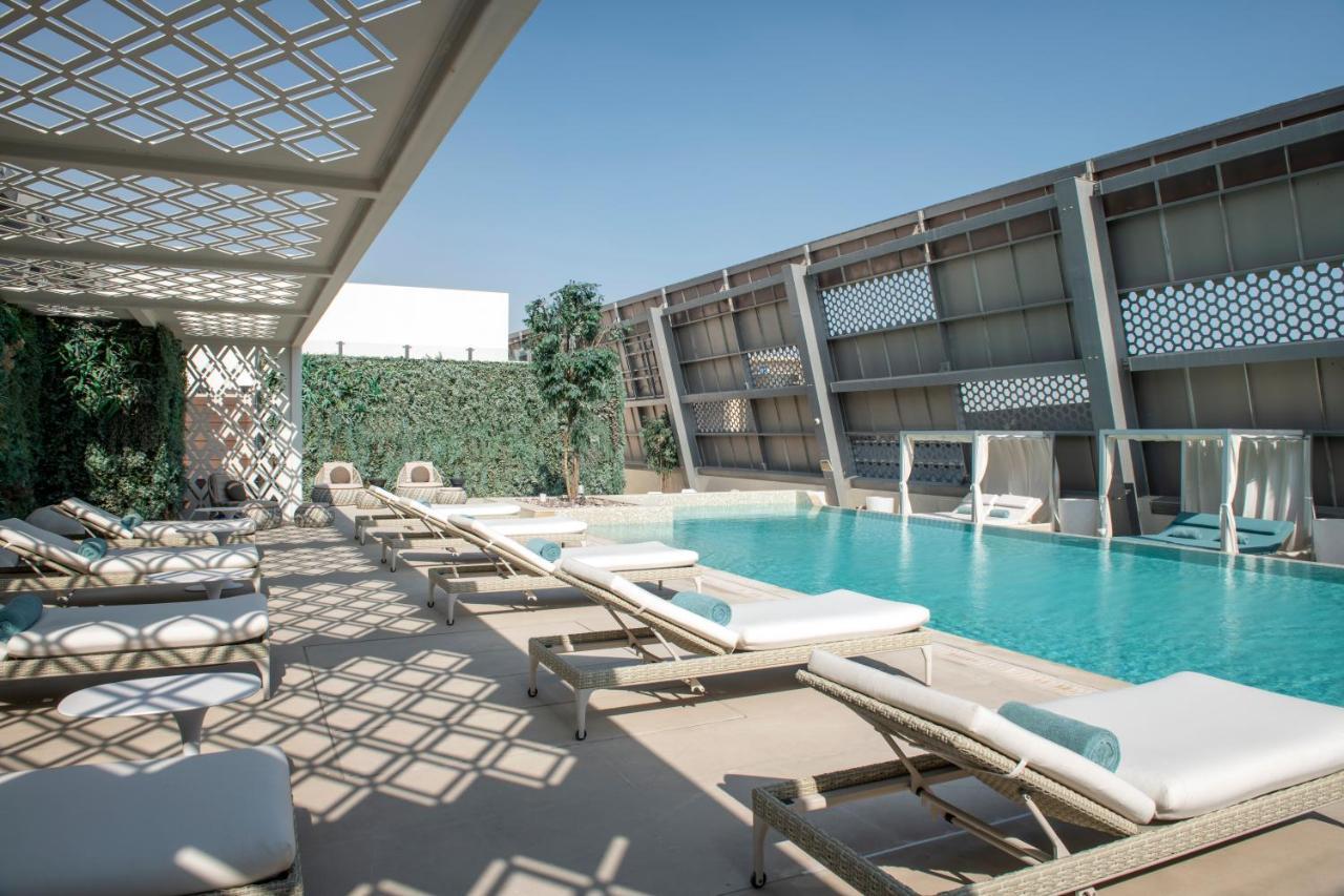 Steigenberger Hotel Doha Exteriör bild The photo shows a modern outdoor pool area. There are several lounge chairs arranged around the pool, which is a vibrant blue color. Sunlight casts intricate shadows on the ground due to a lattice structure overhead. On one side, there are green plan