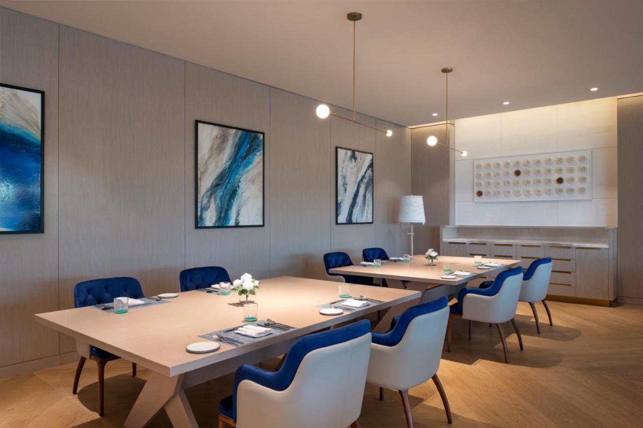 Steigenberger Hotel Doha Exteriör bild The photo shows a modern, elegant dining or meeting room. It features a large, light-colored rectangular table surrounded by chairs with blue upholstery and white frames. Above the table, there are two hanging pendant lights providing illumination. T