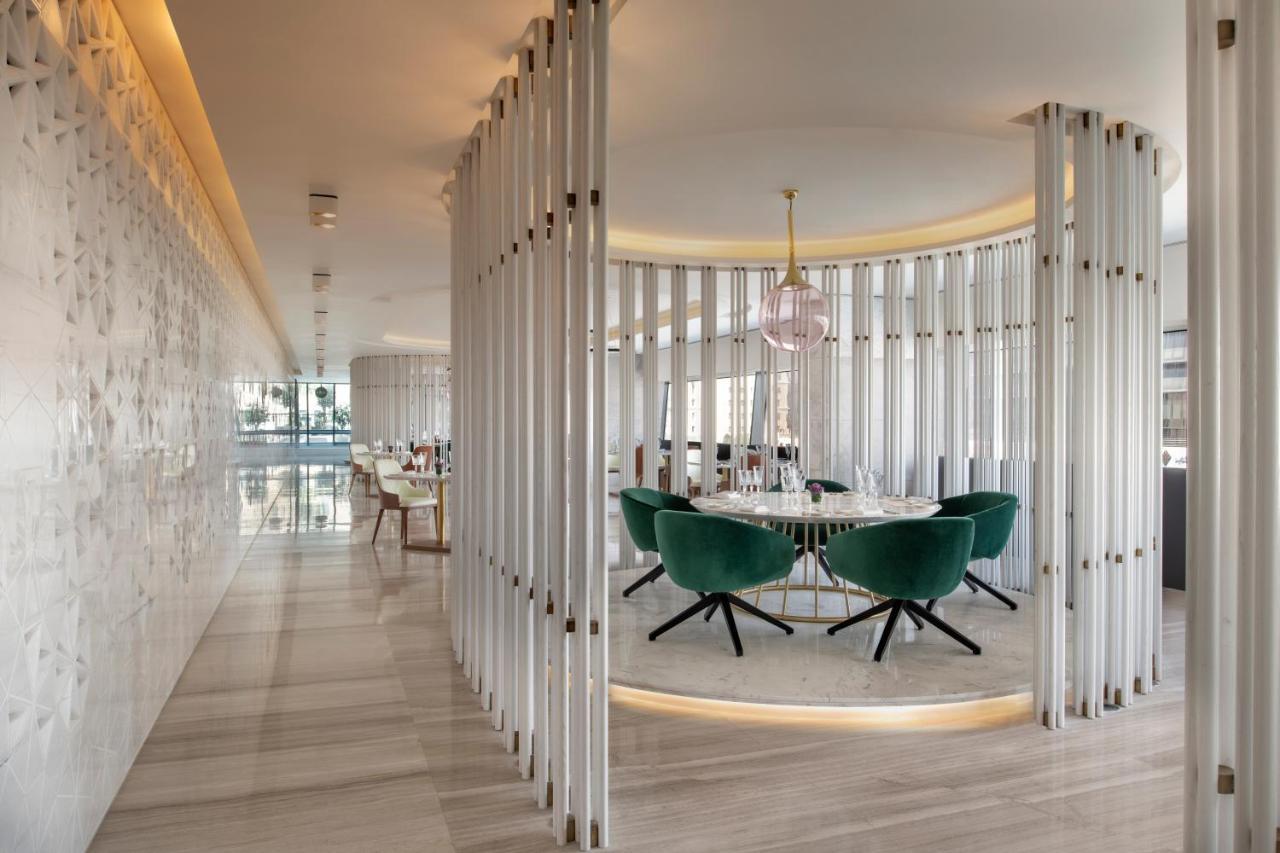 Steigenberger Hotel Doha Exteriör bild The photo shows a modern, stylish interior of a restaurant or dining area. There is a circular dining table surrounded by green upholstered chairs with a sleek design. The space is framed by vertical white panels, creating a sense of separation and p