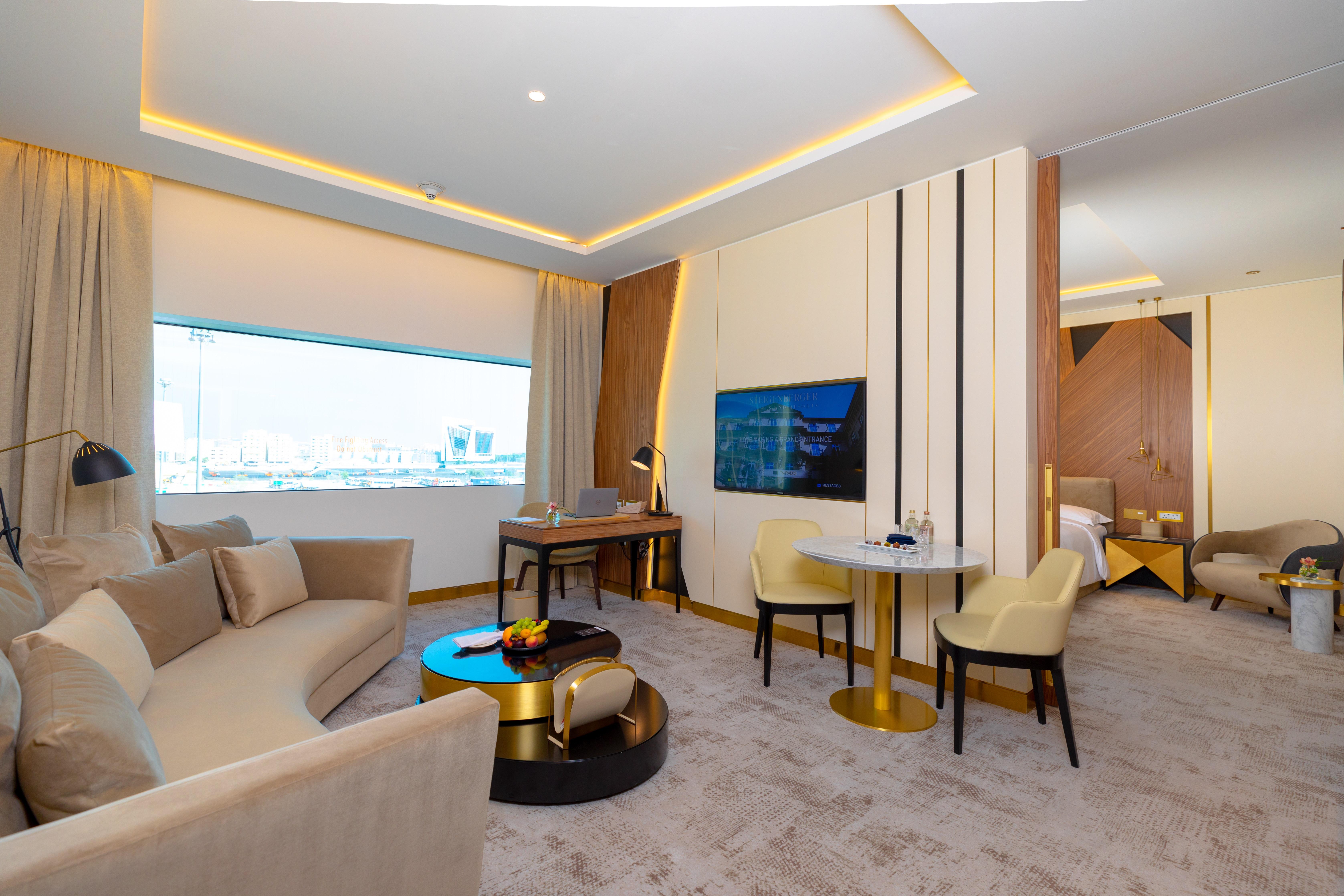 Steigenberger Hotel Doha Exteriör bild The photo shows a modern and stylish hotel suite. It features a spacious living area with a curved sofa set and a small round table accompanied by two chairs. There is a large window that allows natural light to fill the room, enhancing its bright an