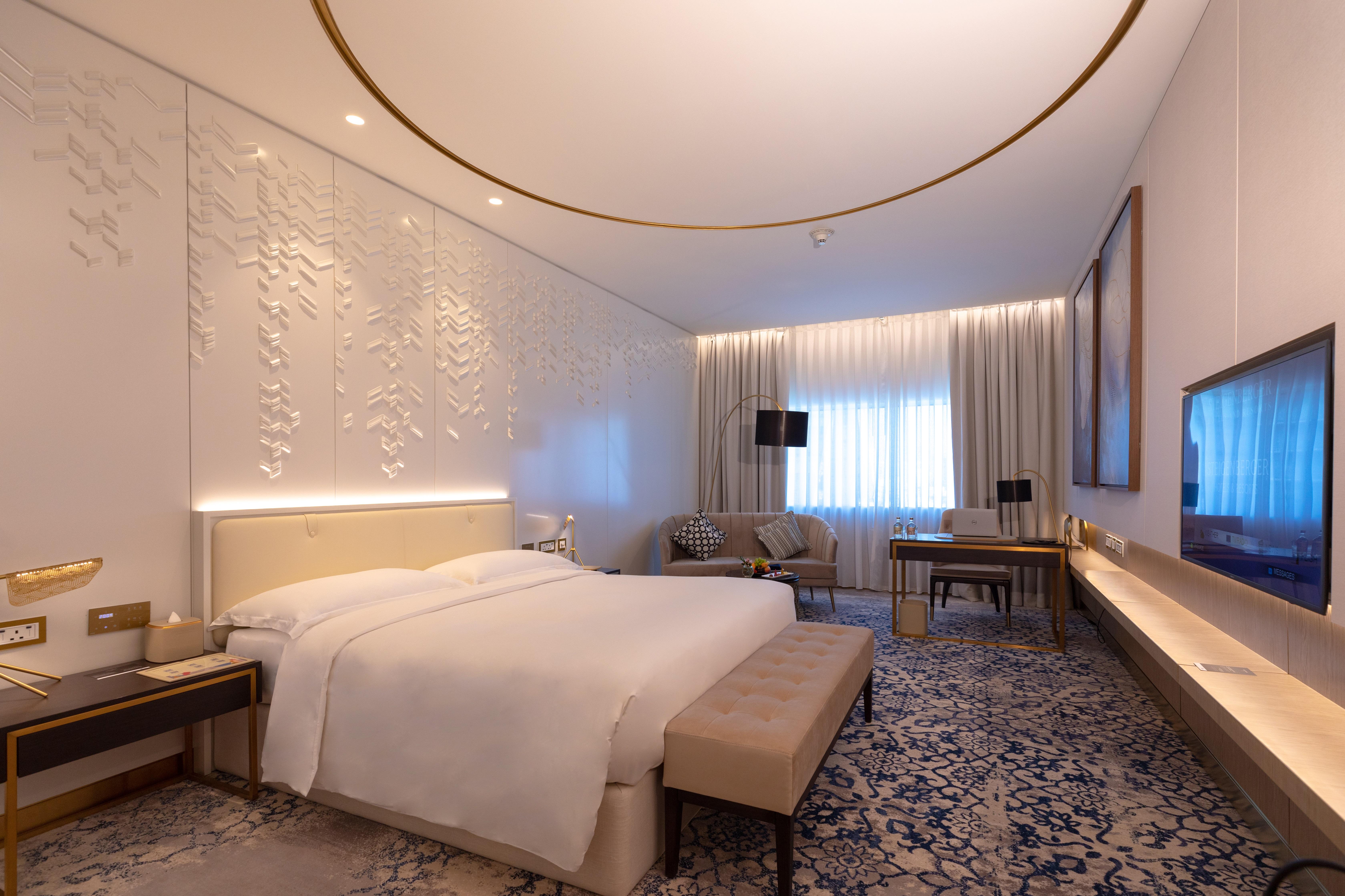 Steigenberger Hotel Doha Exteriör bild The photo shows a modern hotel room designed with a contemporary aesthetic. It features a large bed with white linens, a soft beige bench at the foot of the bed, and elegant furnishings including a bedside table and a comfortable seating area with ch